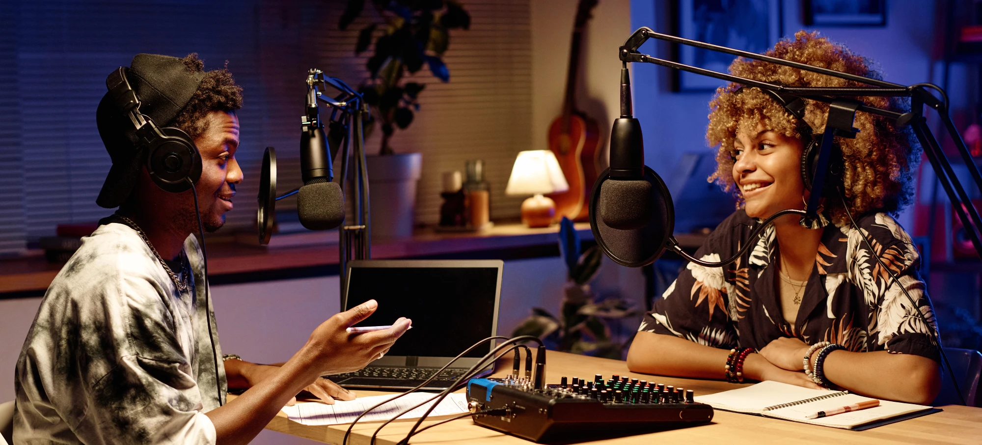 Two people sitting at a table with laptops and microphones.